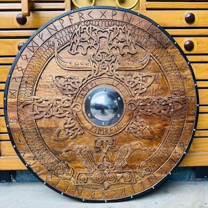 Op de afbeelding: Een rond houten schild met ingewikkelde gravures en een centrale metalen baas. Het schild heeft Noorse ontwerpen, waaronder een boom, raven en knoopwerk, met een donkere rand. Het hout heeft een warme, bruine tint.