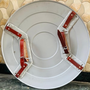 May include: A silver-colored, round shield with a series of concentric circles. The back features two white metal brackets with red leather straps and silver buckles for securing the shield to the arm. The shield has a classic, vintage design.