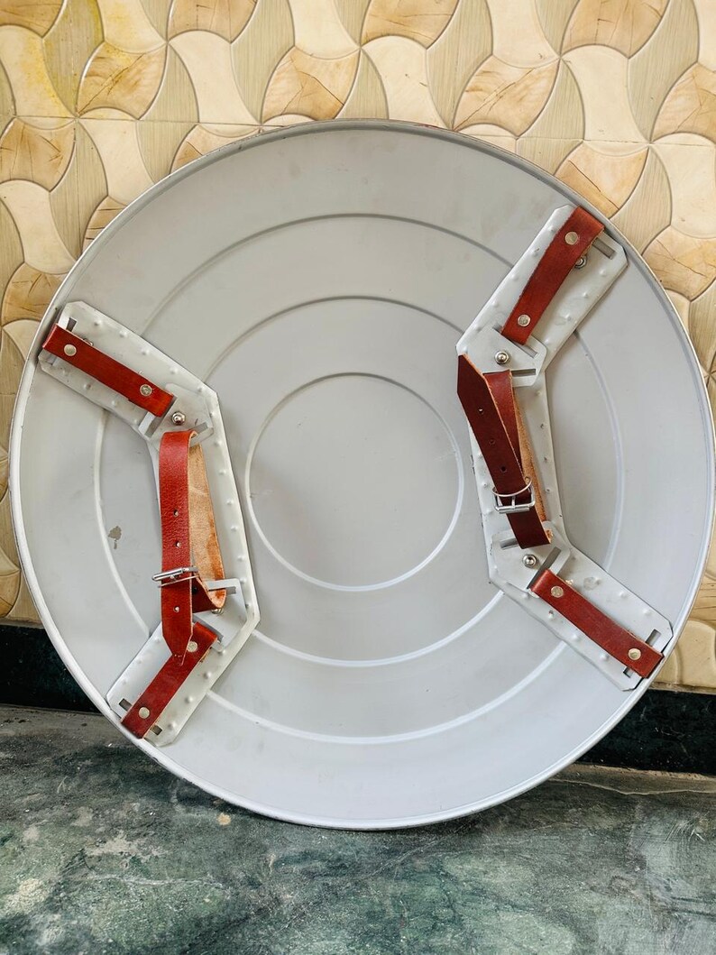 May include: A silver-colored, round shield with a series of concentric circles. The back features two white metal brackets with red leather straps and silver buckles for securing the shield to the arm. The shield has a classic, vintage design.