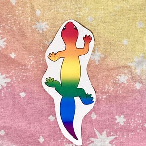 May include: A colorful sticker of a lizard in a rainbow gradient. The lizard has a white outline and is set against a background of pink, yellow, and white fabric with a star pattern. The lizard's colors transition from red to purple.