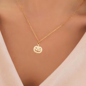 May include: A gold necklace with a small pumpkin pendant. The pendant has carved eyes, nose, and mouth, forming a jack-o'-lantern design. The necklace is worn against a light pink top.