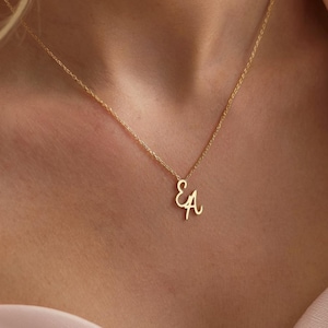 Może przedstawiać: Delikatny złoty naszyjnik z przeplatającymi się literami "E" i "A" jako wisiorek. Naszyjnik jest prezentowany na tle miękkiego różu, podkreślając elegancki design biżuterii. Łańcuszek jest cienki, a wisiorek mały, tworząc minimalistyczną estetykę.