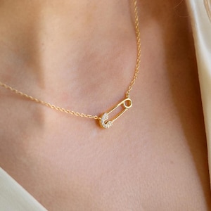 May include: A gold necklace with a safety pin pendant. The safety pin is embellished with small, sparkling stones. The necklace is worn, with the chain and pendant visible against a light background.