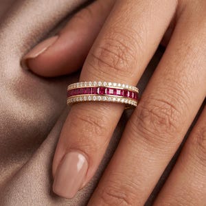 May include: A gold band ring featuring a central row of square-cut deep red rubies flanked by two rows of small, round, clear white diamonds. The ring has a polished metal finish and a channel-set design for the gemstones, creating a continuous band.