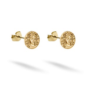 May include: A pair of gold stud earrings. Each earring has a circular design with a detailed relief of a classical female face. The earrings have a post and butterfly backing. The earrings are set against a white background.