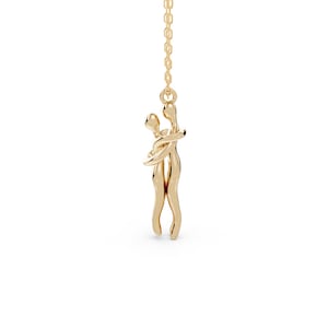 Può includere: Una collana con pendente dorato raffigurante due figure che si abbracciano. Le figure sono intrecciate in un abbraccio, sospese a una catena sottile. Il pendente ha una finitura liscia e lucida, che riflette la luce. La collana è un simbolo di amore e connessione.