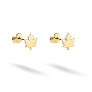 May include: A pair of gold-coloured stud earrings, each shaped like a maple leaf. The earrings are displayed on a white background, with a shadow beneath them.