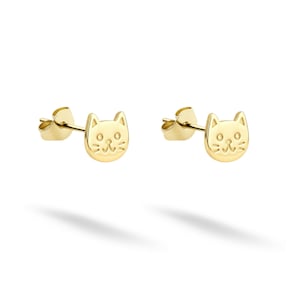 May include: A pair of gold-colored cat-shaped stud earrings. Each earring features a detailed cat face with small ears, eyes, and a nose. The earrings are displayed on a white background, with a shadow beneath them.