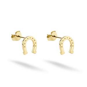 May include: A pair of gold-colored horseshoe stud earrings. Each earring features a horseshoe shape with small, evenly spaced indentations along the curve. The earrings are presented on a white background, with a shadow beneath.