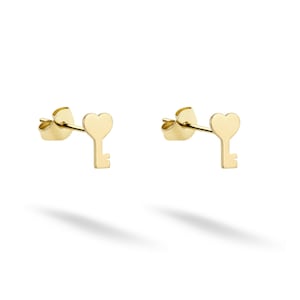 Könnte beinhalten: Ein Paar goldfarbene Ohrstecker. Jeder Ohrring hat eine herzförmige Oberseite und ein schlüsselförmiges Design. Die Ohrringe sind vor einem weißen Hintergrund platziert, mit einem Schatten darunter.