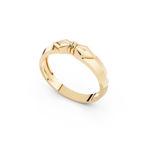 May include: A polished yellow gold ring featuring a central decorative element shaped like two mirrored arrowheads joined by a small double-banded detail. The band has a faceted, angular profile that catches the light, creating a sleek appearance.