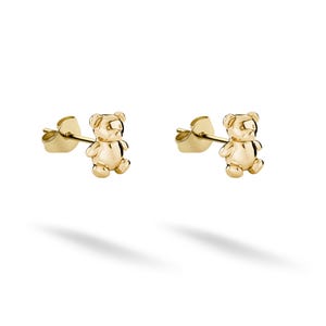 May include: A pair of gold-colored teddy bear stud earrings. Each earring features a small, three-dimensional bear design with detailed features. The earrings are set against a white background, with a shadow beneath.