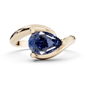 Puede incluir: Un anillo de oro con una gema azul oscuro en forma de pera. La banda se curva alrededor de la piedra, con pequeños detalles dorados. El anillo está sobre un fondo blanco.