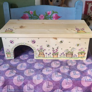 May include: A rectangular light-coloured wooden stool featuring a hand-drawn illustration of potted flowers, bees, and butterflies. The front panel includes an arched cutout opening, and the top surface displays matching floral artwork in the corners.