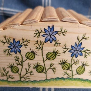 May include: A light-coloured wooden box featuring a hand-drawn illustration of blue flowers with green stems and seed pods. The top of the box has a curved, ribbed design. The base of the front panel is decorated with a green, textured scalloped border.