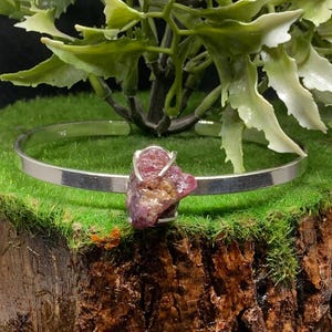 May include: A silver-toned bracelet featuring a raw, reddish-pink gemstone. The stone is held in place by delicate metal prongs. The bracelet is displayed on a mossy surface with greenery in the background, creating a natural aesthetic.