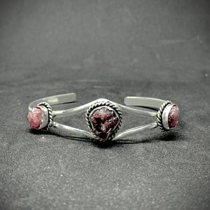 May include: A silver cuff bracelet featuring three raw, reddish-pink gemstones. The bracelet has a simple, elegant design with the stones set in circular bezels. The central stone is larger and framed with a twisted rope design.