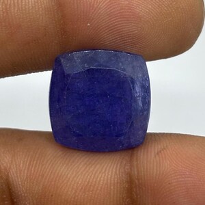 May include: A faceted, cushion-cut gemstone in a deep, saturated blue hue. The gem is held between fingers, showcasing its polished surfaces and the play of light across its facets. The stone's shape is square with rounded corners.