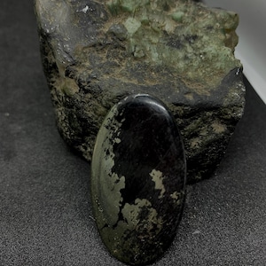 Pode incluir: Uma pedra preta polida em forma oval, com manchas prateadas e douradas, repousando sobre uma superfície escura. Atrás, uma formação rochosa áspera, verde escuro e cinza. A superfície da pedra reflete a luz.