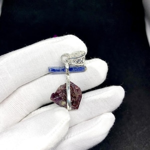 Puede incluir: Un colgante de plata con una gema en bruto de color rojo oscuro. El colgante cuelga de una fianza de plata con un diseño decorativo. Un pequeño cristal azul se coloca encima de la gema. El fondo es oscuro.
