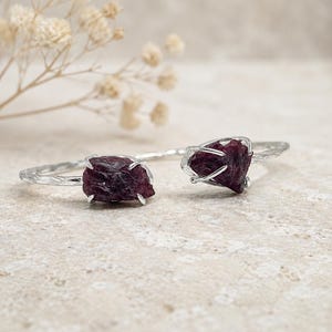May include: A silver-toned cuff bracelet featuring two rough-cut, deep red gemstones. The bracelet has a textured, organic design, with the gemstones held in place by delicate prongs. The background is a neutral, speckled surface.