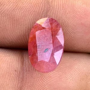 Könnte beinhalten: Oval geschliffener, facettierter Edelstein in einem rötlich-rosa Farbton. Die polierte Oberfläche des Edelsteins weist mehrere Facetten auf, die das Licht reflektieren. Ein kleiner, blau-grüner Einschluss ist im Stein sichtbar. Wahrscheinlich ein Rubin oder ein ähnlicher Edelstein.
