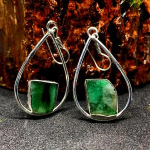 May include: A pair of silver teardrop earrings, each with a vivid green emerald gemstone. The emeralds are set in a polished silver frame, creating a striking contrast. The earrings have a hook closure.