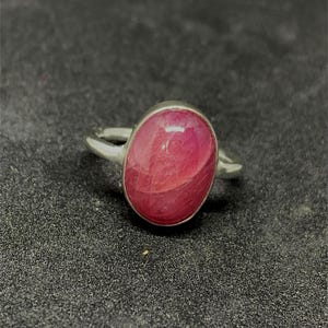 May include: A silver ring with a large, oval-cut ruby gemstone. The ruby is a rich, red hue, set in a plain silver band. The ring is shown against a dark, textured surface. The gemstone is approximately 1.5 cm in length.
