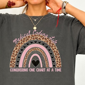 May include: A charcoal grey short-sleeve t-shirt featuring a graphic of a rainbow with leopard print, pink, and black stripes. The text reads Medical Coding Life and Conquering One Chart At A Time, with small black hearts integrated into the design.