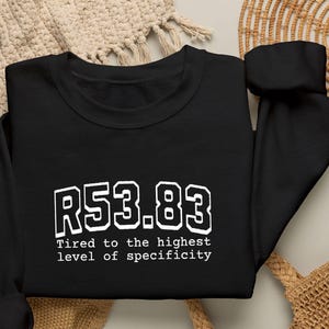 May include: A black long-sleeve sweatshirt featuring white text that reads R53.83 followed by the phrase Tired to the highest level of specificity. The garment has a crew neckline and ribbed cuffs, presenting a clean and straightforward design.