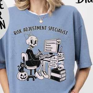 Puede incluir: Una camiseta de color azul vaquero con un gráfico de un esqueleto sentado en un escritorio con el texto "RISK ADJUSTMENT SPECIALIST". El gráfico incluye un monitor de ordenador, un gato negro, una calabaza, una calavera y una nota que dice "Accuracy is my love language".