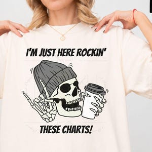 May include: An ivory t-shirt featuring a graphic of a skeleton wearing a grey beanie, holding a coffee cup, and making a rock-and-roll hand gesture. The text reads I'M JUST HERE ROCKIN' THESE CHARTS! in black capital letters above and below the graphic.