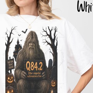 Puede incluir: Una camiseta blanca con un gráfico de un Bigfoot marrón sosteniendo un cartel que dice Q84.2 Other congenital malformations of hair. La escena incluye un bosque oscuro, una casa embrujada, lápidas, murciélagos y calabazas naranjas brillantes.