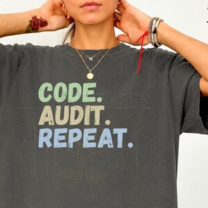 May include: A charcoal gray short-sleeve t-shirt featuring the text CODE. AUDIT. REPEAT. printed in stacked lines. The word CODE is in light green, AUDIT is in tan, and REPEAT. is in light blue, all in a bold, sans-serif font style.