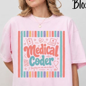 以下が含まれることがあります： 「Medical Coder」および「Decoding care, one code at a time」というテキストが中央にプリントされた、ライトピンクの半袖Tシャツ。クリップボード、ハート、ノートパソコン、本、聴診器のアイコンが描かれ、縦縞の枠で囲まれています。