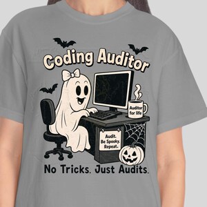 May include: A grey t-shirt featuring a graphic of a ghost sitting at a desk with a computer, a mug labeled 'Auditor for life', and a pumpkin. The text reads 'Coding Auditor' and 'No Tricks. Just Audits.' with a small sign saying 'Audit. Be Spooky. Repeat.'