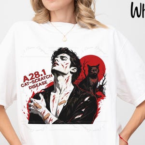 May include: A white t-shirt featuring a graphic print of a person with scratches and bandages, a black cat, and a red sun. The text reads A28.1 CAT-SCRATCH DISEASE. The design includes red and black ink details on a plain white fabric base.