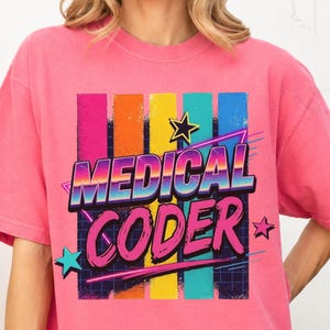 May include: A pink short-sleeve t-shirt featuring the text "MEDICAL CODER" in a bold, multi-colored gradient font. The design includes vertical stripes in pink, orange, yellow, and blue, accented by stars and a grid pattern in the background.