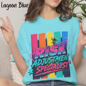May include: A lagoon blue t-shirt featuring a graphic print with vertical stripes in pink, orange, yellow, and teal. The text reads "RISK ADJUSTMENT SPECIALIST" in a stylized font, accented by a star and a pink diagonal line over a grid pattern.