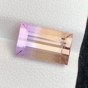 May include: A rectangular gemstone with a gradient of colours. The gem transitions from a light purple hue on one end to a warm, golden-brown shade on the other. The stone is cut with clean, straight lines and facets, reflecting light.