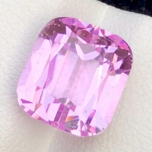 May include: A faceted, rectangular-cut, light pink gemstone. The gem has a slightly rounded shape with multiple facets that reflect light. The stone is set against a white background, highlighting its color and clarity. This gemstone could be used in jewelry.