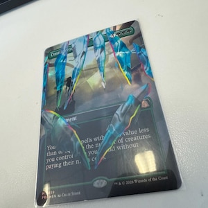 以下が含まれることがあります： 「Omnipresence」と題された長方形のMagic: The Gatheringトレーディングカード。青い結晶の破片のホログラフィックイラストが描かれています。カードには緑のマナシンボル、ゲームテキスト、2026 Wizards of the Coastの著作権表示、アーティスト名が記載されています。