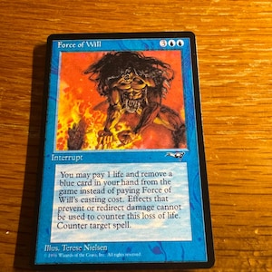 May include: A blue Magic: The Gathering trading card titled Force of Will featuring an illustration of a dark-haired figure amidst flames. The card text reads: You may pay 1 life and remove a blue card in your hand from the game instead of paying Force of Will's casting cost. Effects that prevent or redirect damage cannot be used to counter this loss of life. Counter target spell.