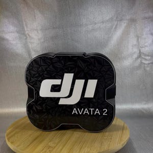 Op de afbeelding: Zwart-witte DJI Avata 2-koffer met een geometrisch patroon. De koffer staat op een houten standaard tegen een zilveren achtergrond. Het DJI-logo en "AVATA 2" worden in het wit weergegeven.