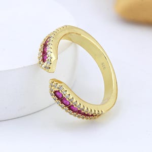 May include: A gold-toned open ring featuring two tapered ends, each inlaid with a row of vibrant red stones bordered by small clear white stones. The inner band is clearly stamped with the number 925, indicating its sterling silver composition.