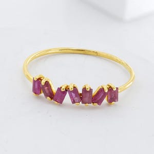 May include: A delicate gold ring featuring a row of vibrant, rectangular ruby gemstones. The ring's slender band and the arrangement of the rubies create a modern and elegant design. The rubies are a deep, rich pink color.