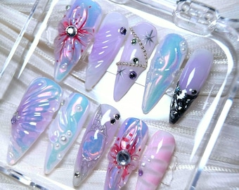 May include: A set of ten iridescent acrylic nails in various designs. The nails feature a range of colors, including pink, purple, and blue, with decorative elements such as flowers, gems, and swirls. The nails are displayed in a clear, rectangular container.