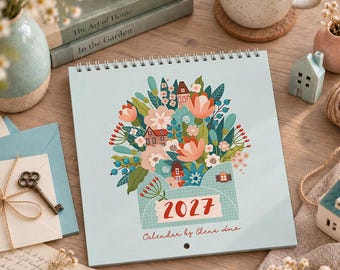 May include: A square 2027 wall calendar with a light blue cover featuring a colorful illustration of a floral bouquet emerging from an envelope, interspersed with small houses. The text 2027 is printed in red on a white label.
