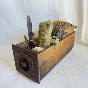 May include: A rustic wooden box with a decorative front panel, filled with a green fabric doll, feathers, a pincushion, and lavender. The doll has a button eye and holds a small bunch of lavender.