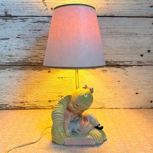 May include: A table lamp featuring a ceramic base shaped like a blonde child sitting in a yellow textured chair while holding a small gray animal. The lamp has a conical, light-colored fabric shade that emits a warm, soft yellow glow.